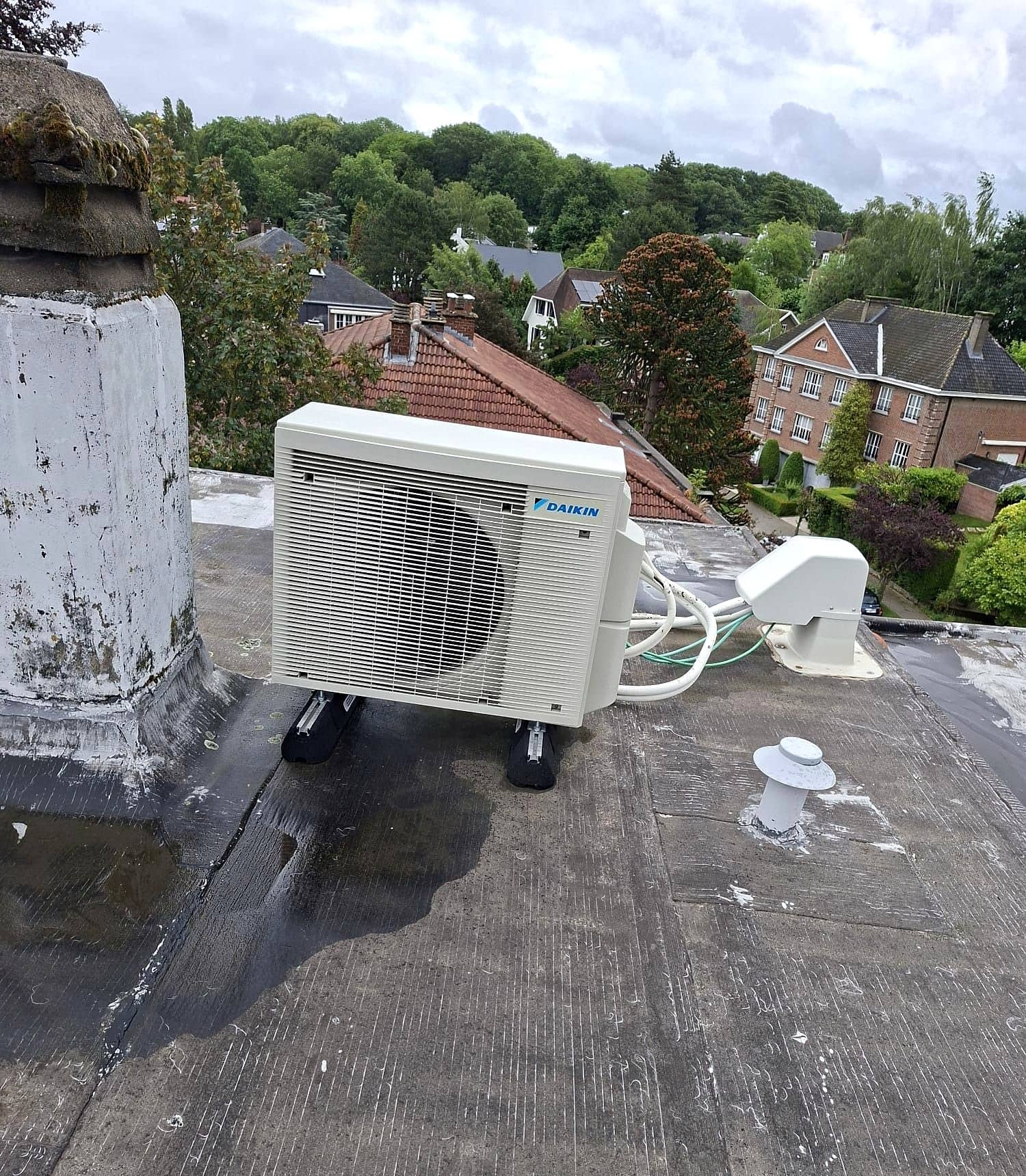 Groupe extérieur Daikin posé sur supports sur un toit plat, avec ses liaisons frigorifiques sous goulotte, maisons et arbres en arrière-plan